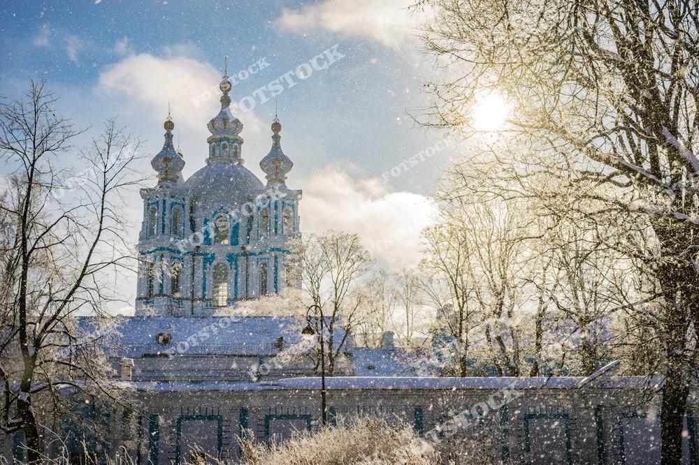 На снимке изображен Смольный собор, окутанный зимней красотой. Солнечный свет, пробивающийся сквозь снежные ветви деревьев, подчеркивает великолепие архитектуры и создает сказочную атмосферу. Собор окружен заснеженным парком, который добавляет уюта и спокойствия. Падающий снег усиливает ощущение зимнего чуда.